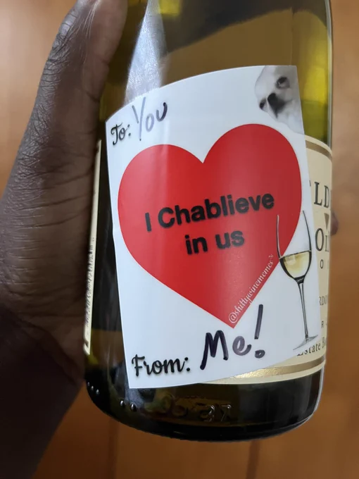Upclose of a dark brown hand holding a wine bottle. The sticker on the wine bottle says To: You, From: Me! “I Chablieve in us” and @shittywinememes in a red heart. A white wine glass and a little white dog on the sticker.