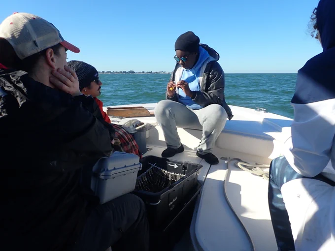 Jasmin shows her crew how to do a shark workup before they haul their first longline. She shits on the bow of the boat with some equipment beside her and the crew sits in a semi circle around her