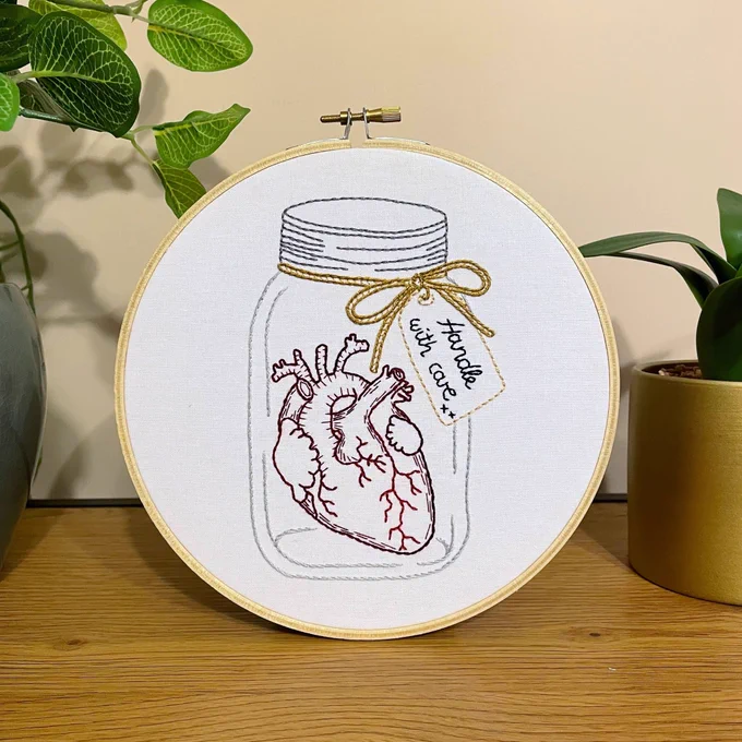An embroidery hoop with the outline of a jar with lid. Inside is an image of an anatomical heart. There is a label attached to the jar, reading “handle with care”