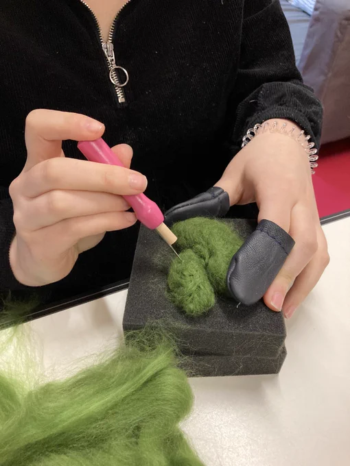 A green needle felted cactus lying flat on a foam block. It is being held by one hand wearing protective black finger pads. The other hand is holding a needle with a pink handle.