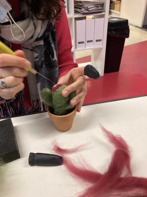 Two hands holding a green needle felted cactus in a terracotta pot. One hand is holding the cactus and Yh w other is holding a needle stabbing the wool to create the felted effect. The pot is on a white table and in the background you can see the red floor.