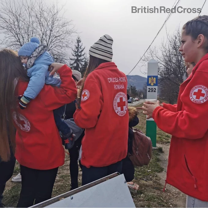 Photograph of three Red Cross volunteers in red uniform, They are with a group of children and the one on the left is holding a baby wearing a blue hat and coat.