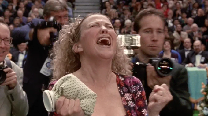 Catherine O'Hara in Best in Show. She is screaming into the air above her, surrounded by photographers and an audience watching the dog show.
