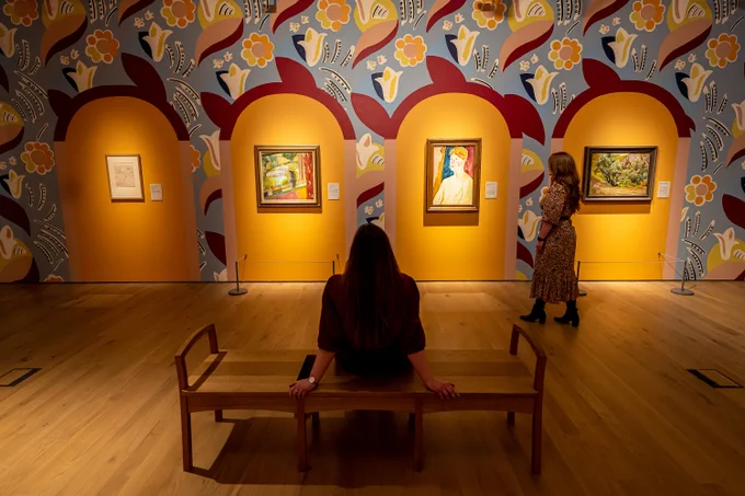 a woman sits on a bench in front of a gallery wall. The wall has bright coloured mural painted on it with flowers. Four artworks hang on the wall. Another woman walks across the space. 