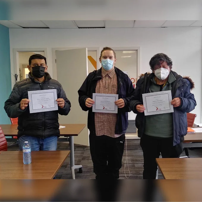3 people stand in a training room. They are all wearing masks, winter clothing and holding up certificates. In front and behind them are brown tables with scattered papers, pens and water bottles on them.