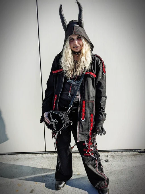 A photo of androidpriest wearing a matching black and red hoodie and pants, accessorized with a coffin bag and demon horns against a white wall background