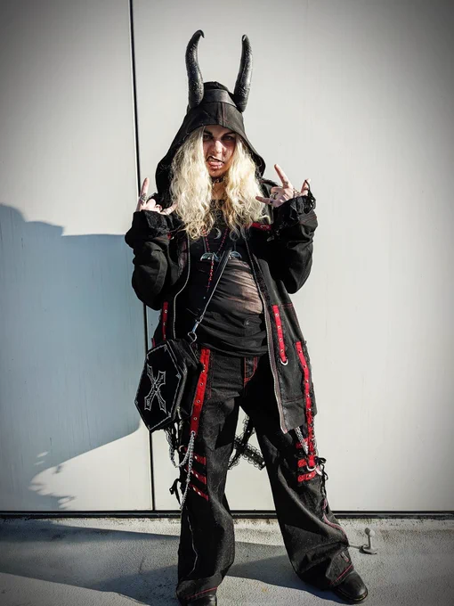 A photo of androidpriest wearing a matching black and red hoodie and pants, accessorized with a coffin bag and demon horns against a white wall background, throwing the horns hand gesture and making a face