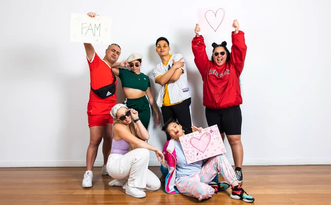 Image Credit: Anna Kucera

Image Description: Members of The Pioneers are posing together in front of a white wall. Four stand in the back with two of them are holding A3 signs. One sign has a big heart on it and the other one has the words 'FAM' written on it. In front of the four people two young women crouch in front of them. One is holding a sign with a coloured in heart.