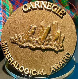 Round gold medal with Carnegie Mineralogical Award written around the edge and a bas relief of a crystal cluster in the middle. It sits on a plexiglass mount in front of some plaid fabric. 