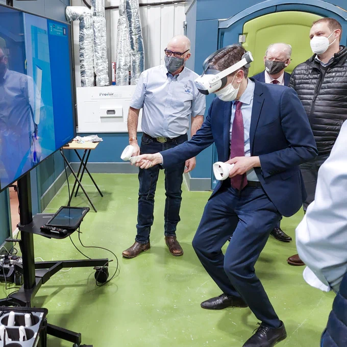 Minister Harris, knees bent, holds a virtual reality controller in each hand, while wearing a VR helmet. In front of him is a large TV screen, and behind the minister are a group watching the screen.