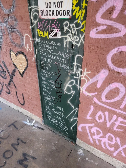 a photo of graffiti on a door at Normandale Park in Portland, OR, where a vigil for June Knightly was held. The graffiti reads "T-Rex was an extremely compassionate comrade and an exemplary ally. They showed up, did the work, and loved on us hard and went home. T-Rex will be missed and loved forever. "