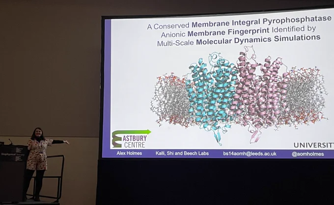 a photo of alex posing dramatically next to the introduction slide to her presentation  which has the title “a conserved membrane integral pyrophosphatase anionic membrane fingerprint identified by multi scale molecular dynamics simulations”