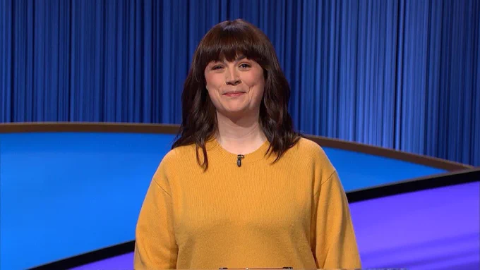 A photograph of Christie, a mid-30s white woman with shoulder length brown hair, smiling and wearing a yellow sweater with a lavier mic clipped on the collar. She is standing in front of the familiar blue Jeopardy set.