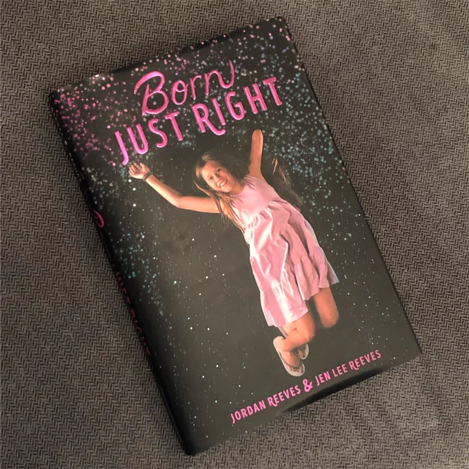 A copy of the Born Just Right book sits on a dark grey tablecloth. The black cover with speckles says “Born Just Right” in purple cursive. There’s a young Jordan jumping with her arms up wearing a purple dress and flip flops. The names “Jordan Reeves & Jen Lee Reeves” are at the bottom.