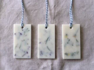 Three rectangular wax shapes, laid out on a sheet of cotton. Each rectangle has a hole in the top centre, which has been threaded with blue and white string.