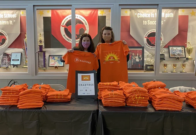 2 female United Arts staff members holing up orange Pieces of Gold tshirts behind a table stacked with tshirts
