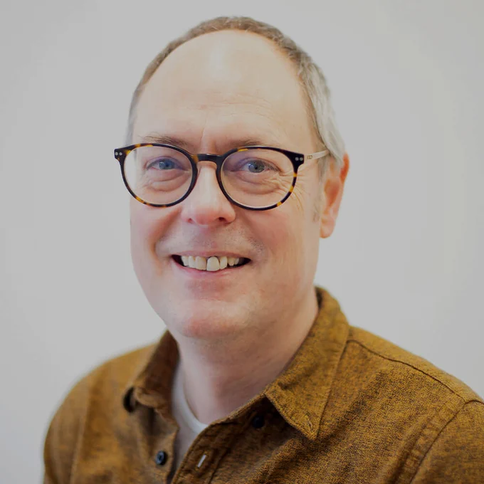 A man wearing glasses and a mustard coloured shirt looks at the camera and smiles