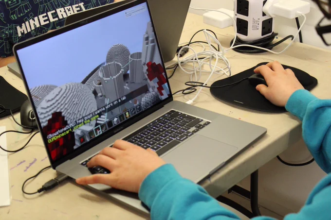 A laptop is on a table with a mouse plugged into it. The mouse is on the right and it is on a black mouse mat. There is a person holding the keyboard and mouse, they are wearing a blue jumper. You can only see their arms. In the background you can see someone’s T-shirt which reads ‘Minecraft’. There is also some cables plugged into an extension cable.