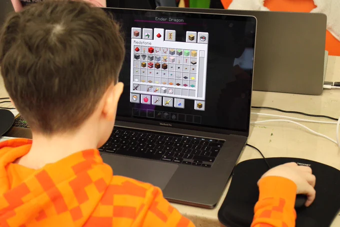 A young boy is sat with his back to the camera. He has brown hair and is wearing an orange square patterned hoody. He sat to a table with a MacBook in front of him playing Minecraft. He is holding a mouse next to the laptop which is on a mouse mat.