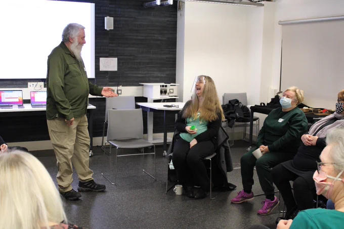 A man is stood facing a group of people in a room with dark flooring and projector screen behind them. He is wearing a dark green shirt with green cargo trousers and boots. He has white hair and beard and is facing a women sat down to his left with his arm out. The woman is wearing a face shield, a blue top and black cardigan and trousers. She is holding a squeeky toy and is laughing.