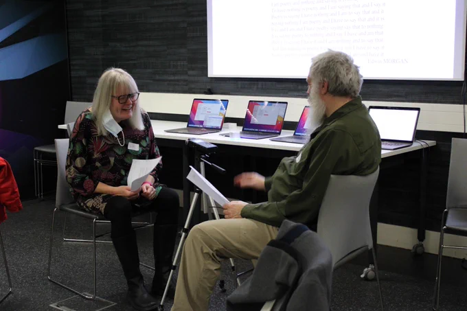 Two people are sat facing eacother with a microphone In between them. The person on the right is the same person stood up in the previous photo. He is holding a piece of paper. The woman opposite him is wearing a floral print top and black trousers and has blonde hair with a fringe. She is also wearing glasses and a face mask is hanging down from her ear. She is laughing and holding a piece of paper. In the background there are MacBooks lined up on the tables and a projector screen above them.