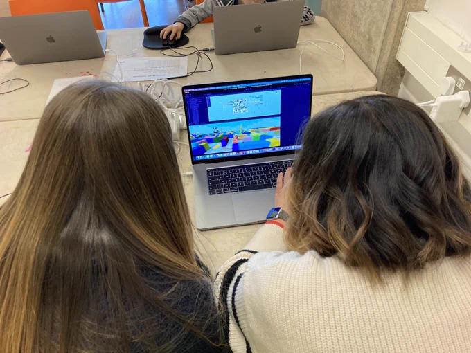 A daughter and parent are sat facing away from the camera at a table with a MacBook on. They are playing a game in a software called ‘Unity’. In the background there are two laptops and one hand is on a mouse next to them.