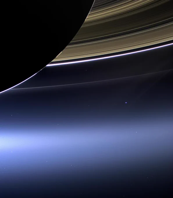 Image of Saturn backlit by the Sun. A quarter of its large rings are at the top right and the image is overexposed to show the uniilluminated sphere of Saturn. Below the rings is a tiny star-looking dot, which is Earth.