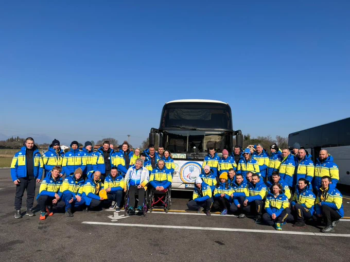 A picture of many athletes dressed in blue and yellow clothing posing in front of a bus featuring the NPC Ukraine logo. The sky is blue with no clouds.