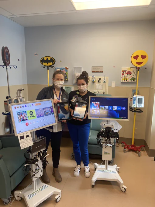 Two child life specialists holding iPads and standing between two mobile gaming carts in a paediatric hospital treatment room.