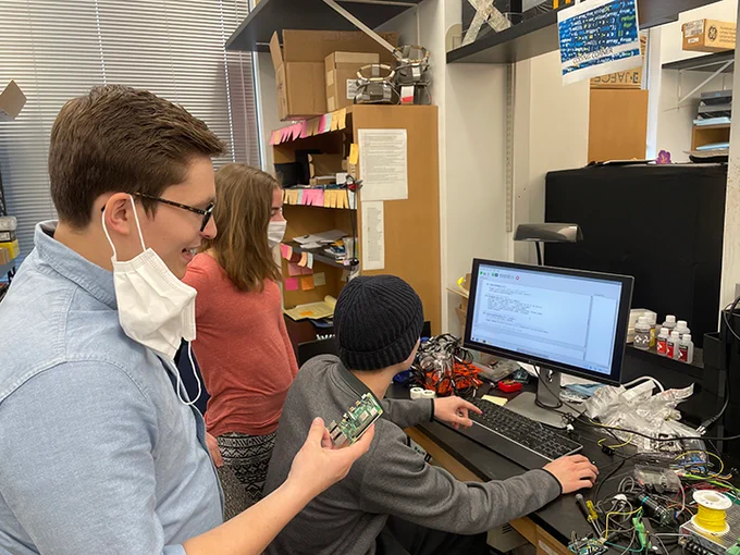 Landon is in a blue shirt holding a green circuit. Drew is in front of Landon and is sitting in front of a computer with code on the screen. Emily is in a pink sweater looking at the screen to the left of Drew.