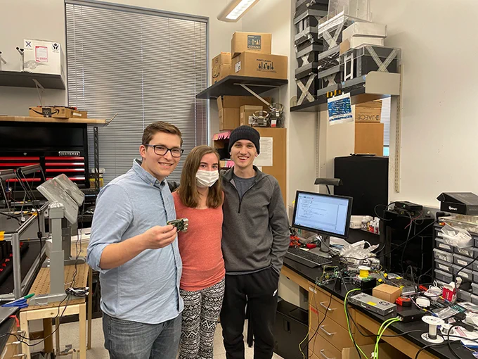 From the left to the right, Landon Emily and Drew are standing together in a lab. The room is filled with circuits, papers and robotic equipment.