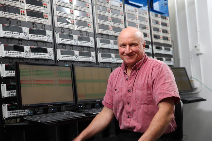 A mature, balding man wearing a short-sleeved button-down shirt sits in front of three computer screens and keyboards. He is smiling.