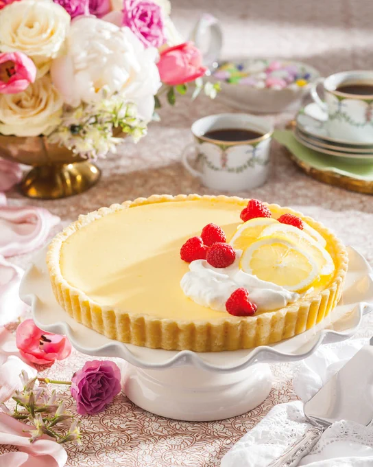 Southern Lady’s Lemon Raspberry Tart displayed on a white ruffle-edged cake stand, garnished with whipped cream, fresh raspberries, and lemon slices.