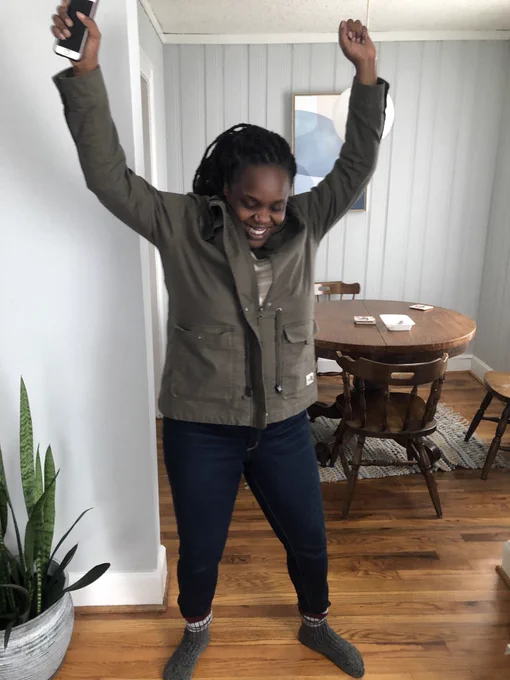 A Black woman with both of her arms in their air with a joyous look of victory.  She is in her dining room with a table and snake plant behind her.