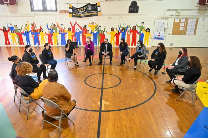 Michelle, MA state leaders, and school staff sit in a circle inside a school gymnasium with a colorful mural depicting students in the background.