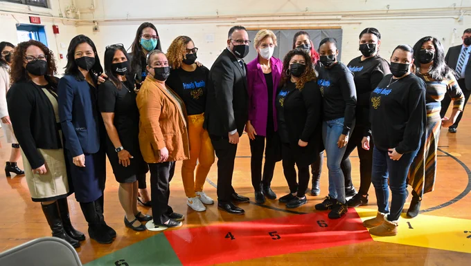 Michelle, MA state leaders, and school staff pose for a group photo inside a sunny gymnasium.