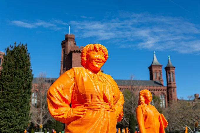 Orange statue of woman with hands on hips. Behind her, another state holding something in hand. In background, Smithsonian Castle.