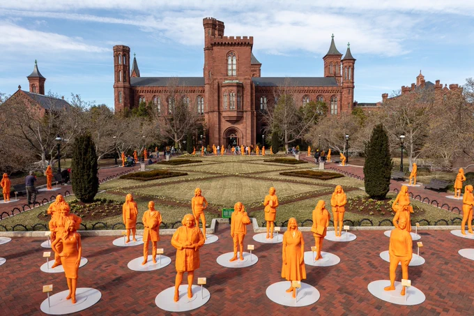 More than a dozen bright orange statues of women scientists on display in a garden on a sunny day. In background, Smithsonian Castle. 