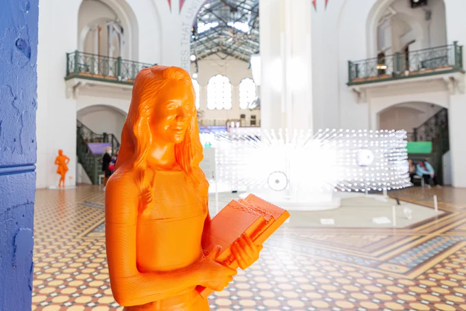 Orange statue of a woman holding books or notepads in the courtyard of a museum building. Behind her, a light-up art installation.