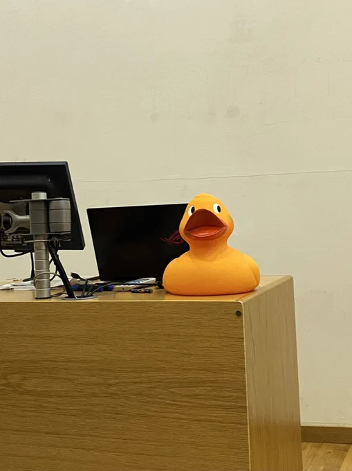 A large yellow plastic duck on top of a wooden desk. The desk has a black laptop and monitor on it.
