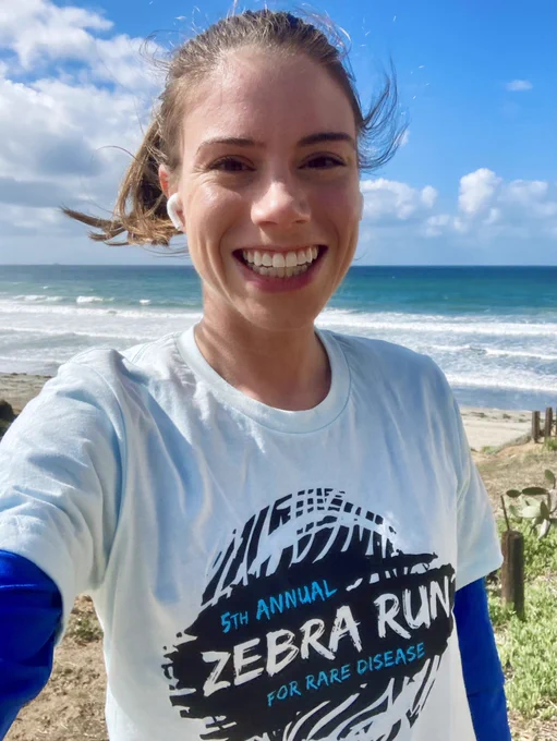 Selfie of Naomi standing by the ocean. Naomi is wearing a light blue shirt that says “5th Annual Zebra Run for Rare Diseases.” Naomi’s brown hair is windswept.