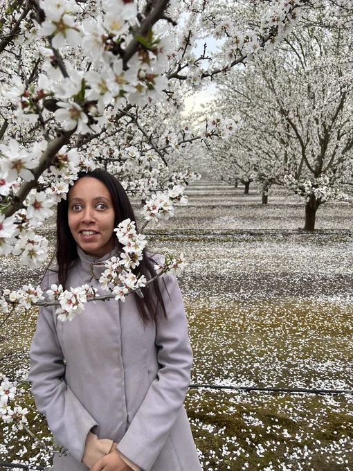 I’m a Black disabled woman smiling in an orchard. Small flowers cover the trees and the ground is dotted with white petals. Almond blossms infuse the air with the sweet but delicate fragrance of hope.