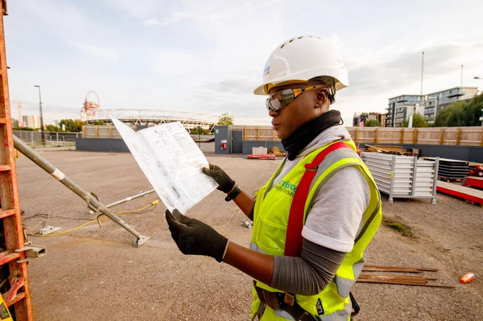 Woman in high-viz and hard hat reads plans, building site in background.