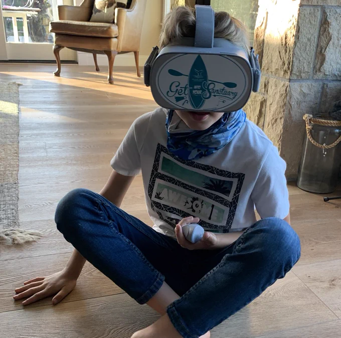 A young child sits cross-legged on the floor with Oculus Go goggles on the child's head and controller in hand. The sticker on the goggles reads “Get Into Your Sanctuary”. 
Credit: Claire Fackler/NOAA National Marine Sanctuaries