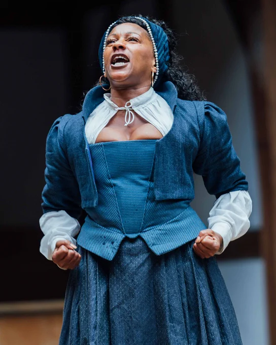 An actor wearing a blue Elizabethan corset gown defiantly stands on stage, their arms at waist height, fists clenched, and they fiercely deliver a speech.