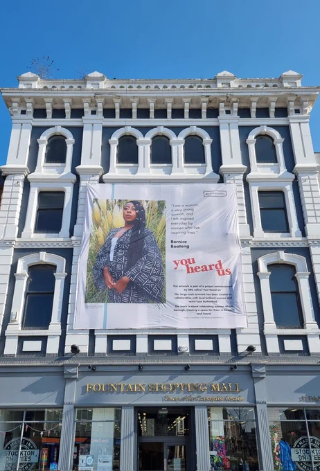 A huge banner showing a portrait of local woman Bernice Boateng, and carrying info about the You Heard Us project, hangs in the sunshine on the exterior of the Fountain Shopping Mall.