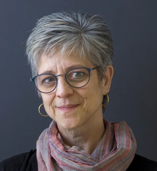 A light source highlights the left side of Dr. Shelley Wall's face. Dr. Wall stands in front of a dark grey background. She has short, spiky salt-and-pepper hair and gamine features. She wears glasses, gold hoop earrings and has a pink-and-grey scarf wound around her neck.
