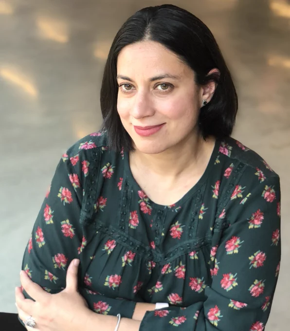 A professional headshot of Saima Sitwat, a Pakistani American woman. She has shoulder-length black hair and hazel eyes. She wears a green shirt with pink and yellow flowers on it. Her arms are crossed. 