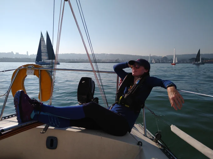 Karissa steering warpath with her feet reclining up against the life lines. Multiple sailboats are in the background sailing in San Francisco bay.