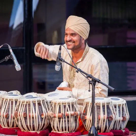 An image of someone sitting on the floor and playing on drums. They are wearing gold clothing and are smiling. Microphones are positioned above the drums.